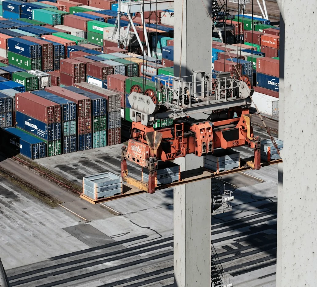 A port filled with containers in various colors. In the foreground, an orange container is lifted