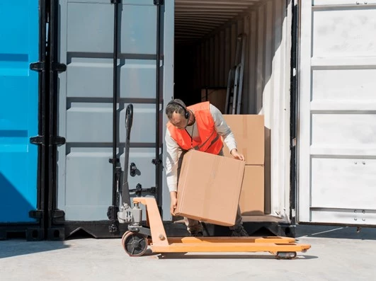 En man som bär in kartonger i en grå container. Mannen är iklädd en orange varselväst och har på sig hörlurar.