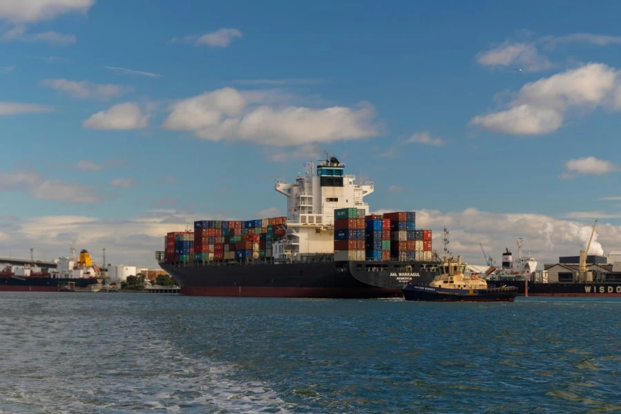Ett stort containerfartyg lastat med färgglada containrar ligger vid kaj, assisterat av en bogserbåt.