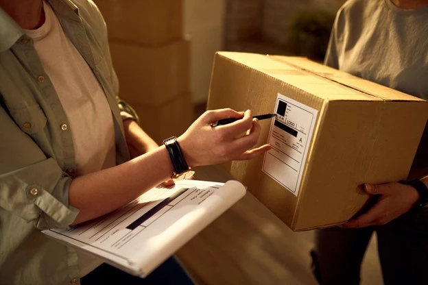 Närbild av två personer som hanterar en paketleverans. En person håller ett paket med en fraktetikett, medan den andra markerar etiketten med en penna och håller en checklista. Kartonger syns i bakgrunden.