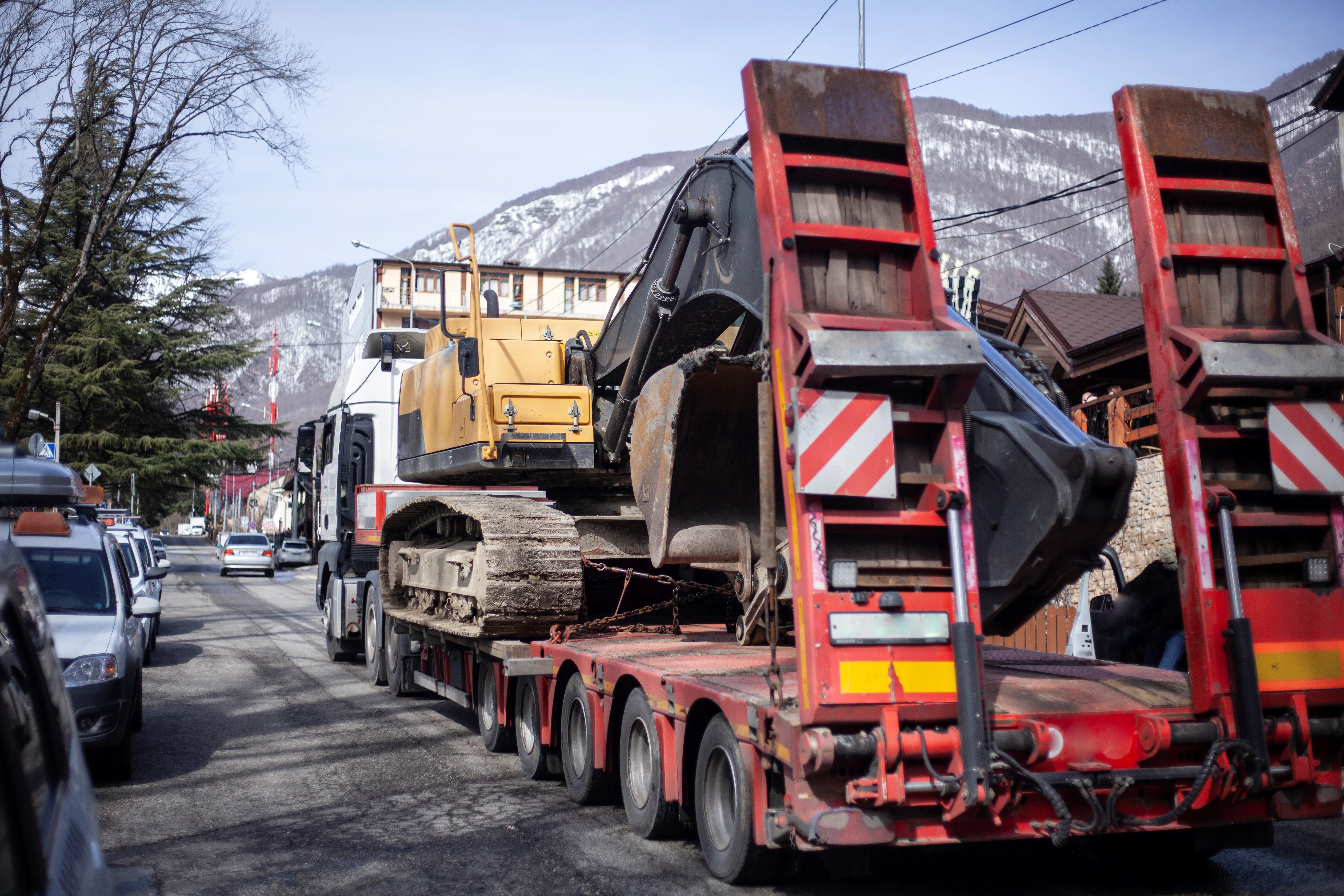a yellow excavator loaded onto a flatbed truck, driving along a narrow street with trees lining the road. The scene is set in a mountainous area, with snow-covered peaks visible in the background. The truck carrying the excavator is positioned between other cars on the street.