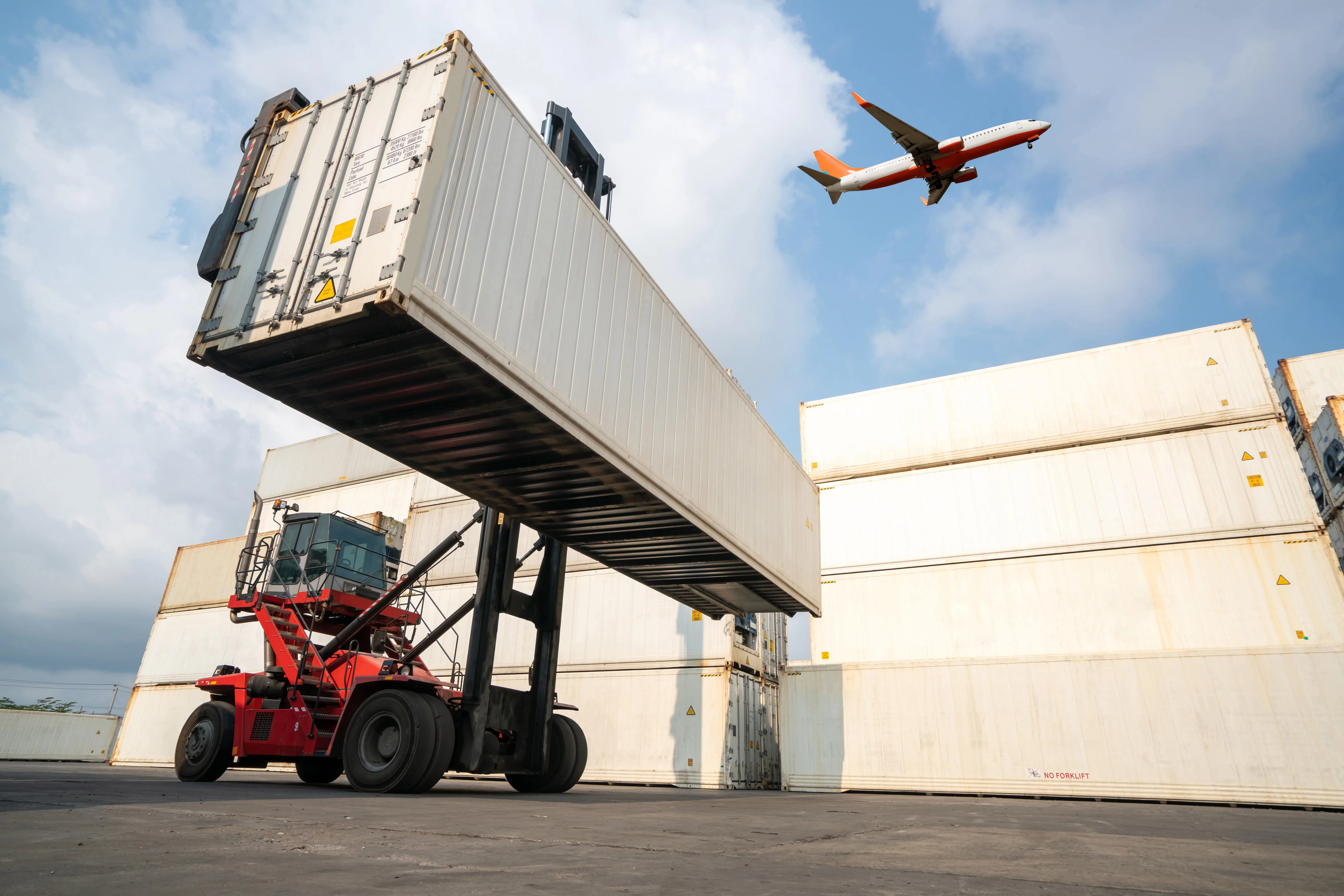 A red forklift lifting a large white shipping container in a freight yard, with stacked containers in the background and a commercial airplane flying overhead against a blue sky.