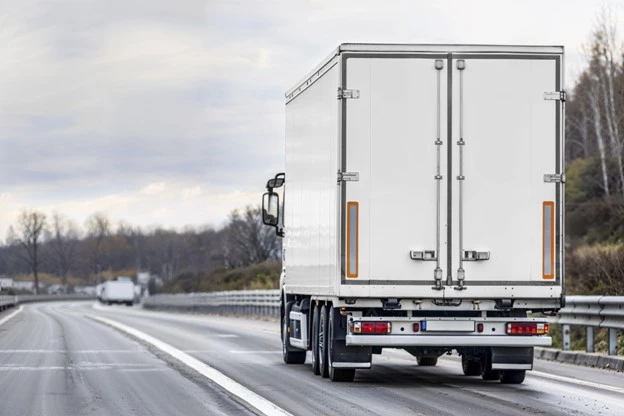 En vit lastbil med en sluten kyl- eller transportcontainer kör längs en motorväg. Vägen är omgiven av träd och ett grått molntäcke syns på himlen. Andra lastbilar syns i bakgrunden.