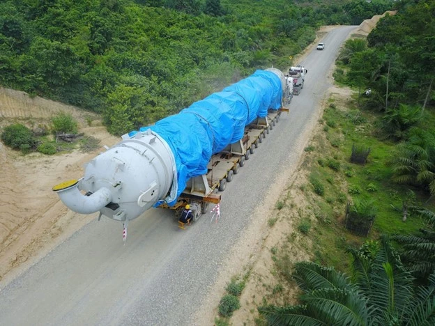 Aerial view of an oversized industrial component covered with a blue tarp being transported on a multi-axle trailer along a rural road surrounded by dense green vegetation and palm trees.