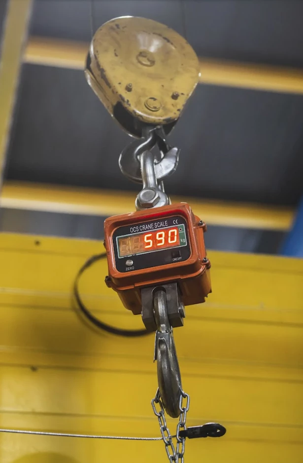 En industriell kranvåg som visar vikten 590 kg, hängande från en krok med kedjor i en lager- eller verkstadsmiljö.