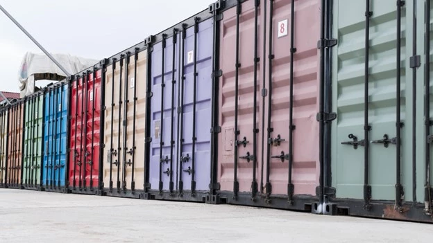 Colorful row of shipping containers in various sizes and shades, lined up outdoors on a concrete surface.